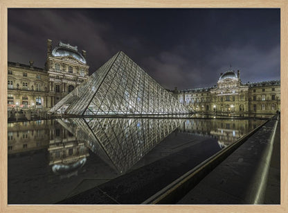 A dramatic night photograph of the illuminated Louvre Pyramid in Paris, its glass structure glowing against a dark, cloudy sky. The historic Louvre Palace is visible in the background, and the entire scene is perfectly reflected in the still water of a foreground pool. The image is presented within a silver frame. Artwork