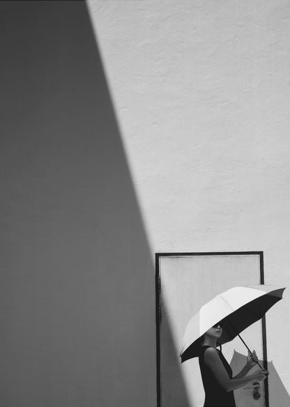 A minimalist black and white photograph of a woman in a black dress holding a two-toned umbrella. She stands in front of a door against a light wall, with a sharp, diagonal shadow dramatically cutting across the upper left of the image, creating a high-contrast, geometric composition. Decor