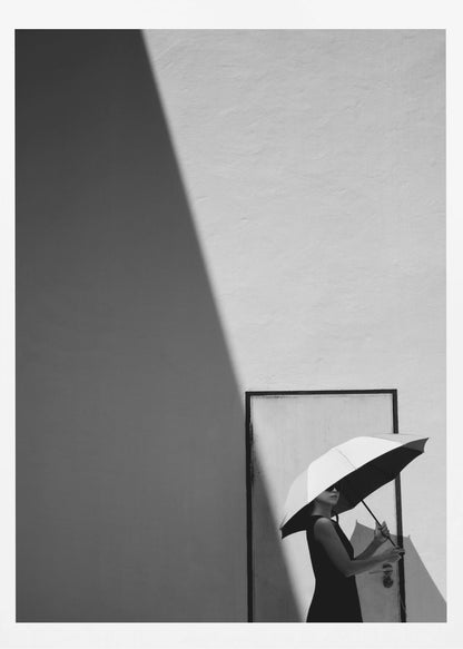 A minimalist black and white photograph of a woman in a black dress holding a two-toned umbrella. She stands in front of a door against a light wall, with a sharp, diagonal shadow dramatically cutting across the upper left of the image, creating a high-contrast, geometric composition. Decor