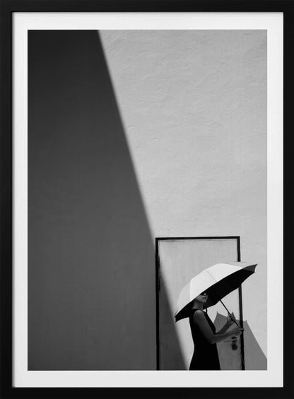 A minimalist black and white photograph of a woman in a black dress holding a two-toned umbrella. She stands in front of a door against a light wall, with a sharp, diagonal shadow dramatically cutting across the upper left of the image, creating a high-contrast, geometric composition. Decor