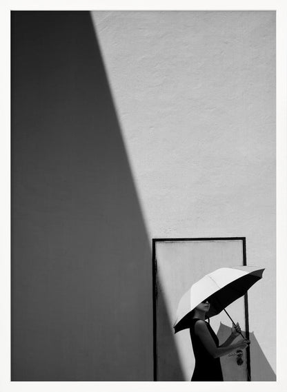 A minimalist black and white photograph of a woman in a black dress holding a two-toned umbrella. She stands in front of a door against a light wall, with a sharp, diagonal shadow dramatically cutting across the upper left of the image, creating a high-contrast, geometric composition. Decor