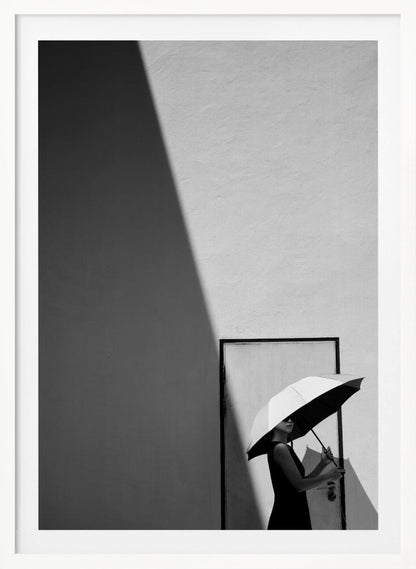 A minimalist black and white photograph of a woman in a black dress holding a two-toned umbrella. She stands in front of a door against a light wall, with a sharp, diagonal shadow dramatically cutting across the upper left of the image, creating a high-contrast, geometric composition. Decor