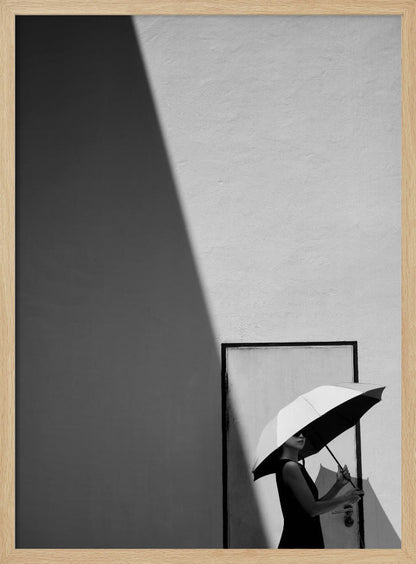 A minimalist black and white photograph of a woman in a black dress holding a two-toned umbrella. She stands in front of a door against a light wall, with a sharp, diagonal shadow dramatically cutting across the upper left of the image, creating a high-contrast, geometric composition. Decor