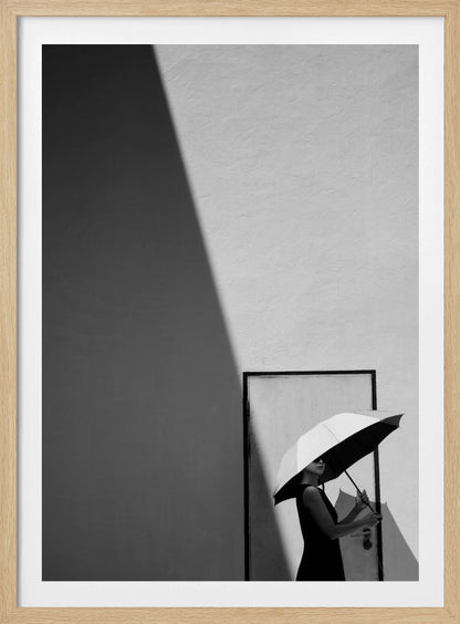 A minimalist black and white photograph of a woman in a black dress holding a two-toned umbrella. She stands in front of a door against a light wall, with a sharp, diagonal shadow dramatically cutting across the upper left of the image, creating a high-contrast, geometric composition. Decor