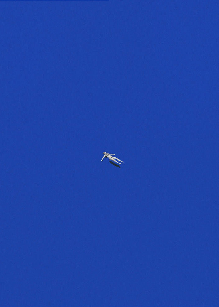 A minimalist and surreal artwork featuring a tiny, pale human figure floating peacefully in the center of an immense, solid, and vibrant blue void, all enclosed within a thin black frame. Wall Art