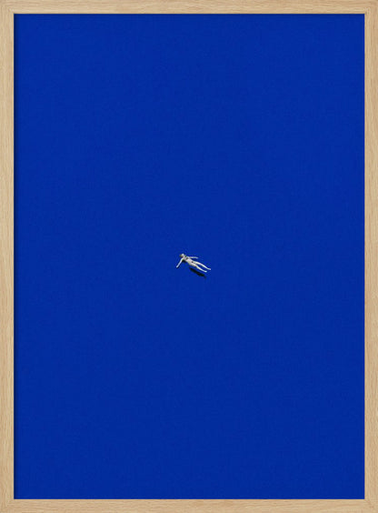A minimalist and surreal artwork featuring a tiny, pale human figure floating peacefully in the center of an immense, solid, and vibrant blue void, all enclosed within a thin black frame. Wall Art