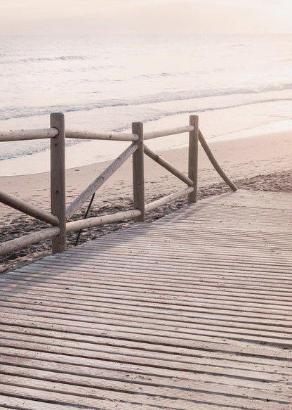 A wooden boardwalk with a rustic log fence leads down to a sandy beach with gentle waves lapping at the shore under a soft, hazy sunrise or sunset. Decor