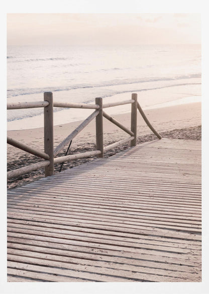 A wooden boardwalk with a rustic log fence leads down to a sandy beach with gentle waves lapping at the shore under a soft, hazy sunrise or sunset. Decor