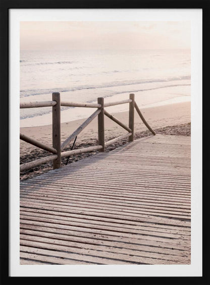 A wooden boardwalk with a rustic log fence leads down to a sandy beach with gentle waves lapping at the shore under a soft, hazy sunrise or sunset. Decor