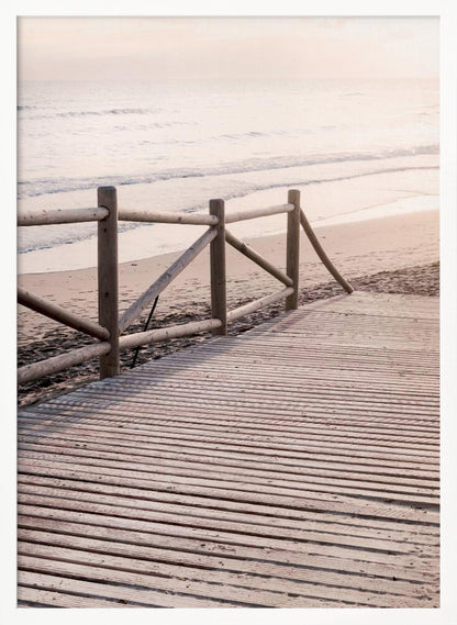 A wooden boardwalk with a rustic log fence leads down to a sandy beach with gentle waves lapping at the shore under a soft, hazy sunrise or sunset. Decor