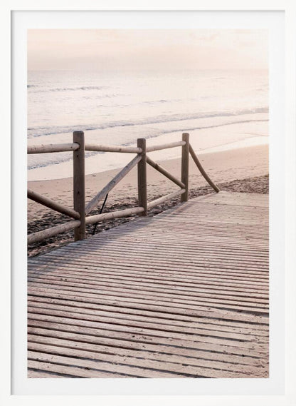 A wooden boardwalk with a rustic log fence leads down to a sandy beach with gentle waves lapping at the shore under a soft, hazy sunrise or sunset. Decor