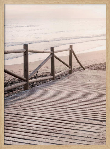 A wooden boardwalk with a rustic log fence leads down to a sandy beach with gentle waves lapping at the shore under a soft, hazy sunrise or sunset. Decor