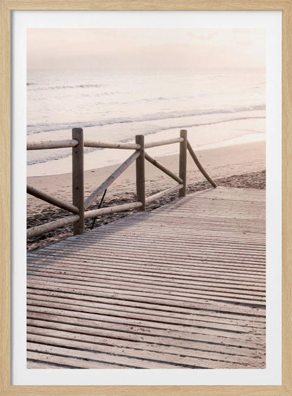 A wooden boardwalk with a rustic log fence leads down to a sandy beach with gentle waves lapping at the shore under a soft, hazy sunrise or sunset. Decor