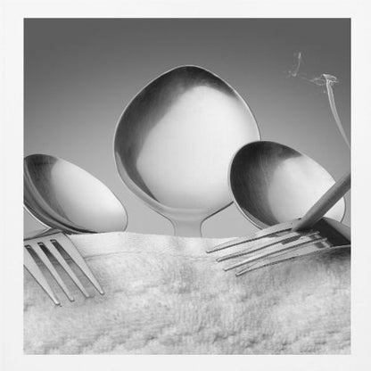 A conceptual black and white photograph in a light wood frame, featuring three spoons standing upright on a white towel, while a fork in the foreground holds a lit, smoking cigarette between its tines. Wall Art