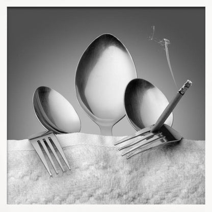 A conceptual black and white photograph in a light wood frame, featuring three spoons standing upright on a white towel, while a fork in the foreground holds a lit, smoking cigarette between its tines. Wall Art
