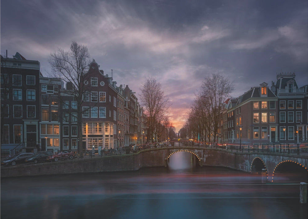 A dramatic long-exposure photograph of an Amsterdam canal at twilight. An arched bridge spans the water, lined by historic Dutch buildings with glowing windows. The sky is filled with dark, moody clouds above a fading orange and purple sunset, and red light trails from boats are blurred on the water's surface. Poster