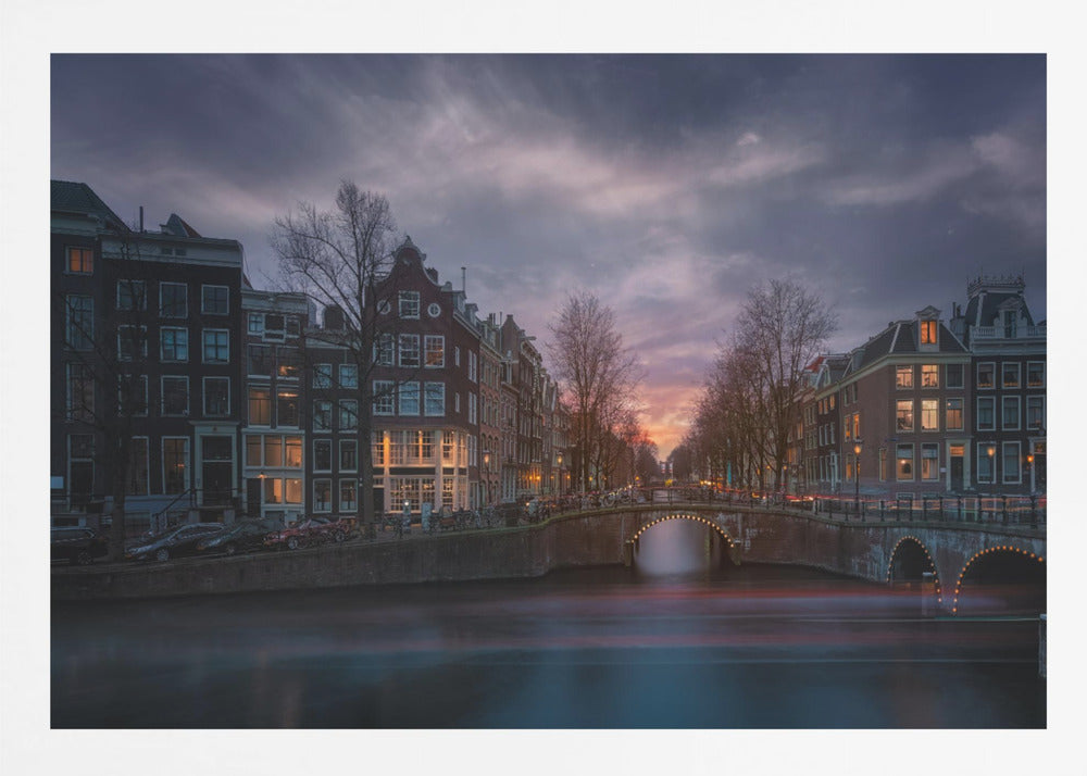 A dramatic long-exposure photograph of an Amsterdam canal at twilight. An arched bridge spans the water, lined by historic Dutch buildings with glowing windows. The sky is filled with dark, moody clouds above a fading orange and purple sunset, and red light trails from boats are blurred on the water's surface. Poster