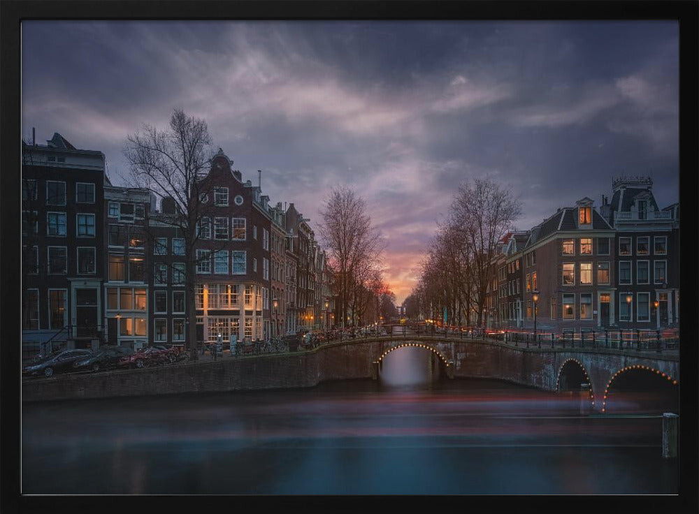 A dramatic long-exposure photograph of an Amsterdam canal at twilight. An arched bridge spans the water, lined by historic Dutch buildings with glowing windows. The sky is filled with dark, moody clouds above a fading orange and purple sunset, and red light trails from boats are blurred on the water's surface. Poster