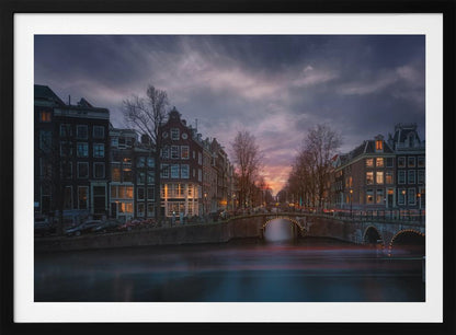 A dramatic long-exposure photograph of an Amsterdam canal at twilight. An arched bridge spans the water, lined by historic Dutch buildings with glowing windows. The sky is filled with dark, moody clouds above a fading orange and purple sunset, and red light trails from boats are blurred on the water's surface. Poster