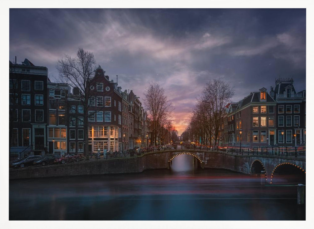 A dramatic long-exposure photograph of an Amsterdam canal at twilight. An arched bridge spans the water, lined by historic Dutch buildings with glowing windows. The sky is filled with dark, moody clouds above a fading orange and purple sunset, and red light trails from boats are blurred on the water's surface. Poster