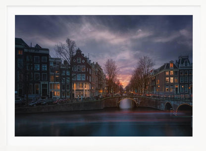 A dramatic long-exposure photograph of an Amsterdam canal at twilight. An arched bridge spans the water, lined by historic Dutch buildings with glowing windows. The sky is filled with dark, moody clouds above a fading orange and purple sunset, and red light trails from boats are blurred on the water's surface. Poster