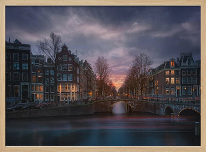 A dramatic long-exposure photograph of an Amsterdam canal at twilight. An arched bridge spans the water, lined by historic Dutch buildings with glowing windows. The sky is filled with dark, moody clouds above a fading orange and purple sunset, and red light trails from boats are blurred on the water's surface. Poster