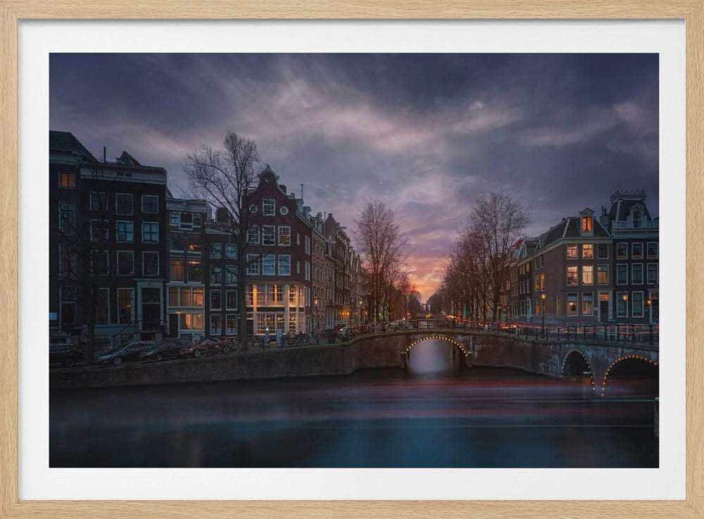 A dramatic long-exposure photograph of an Amsterdam canal at twilight. An arched bridge spans the water, lined by historic Dutch buildings with glowing windows. The sky is filled with dark, moody clouds above a fading orange and purple sunset, and red light trails from boats are blurred on the water's surface. Poster