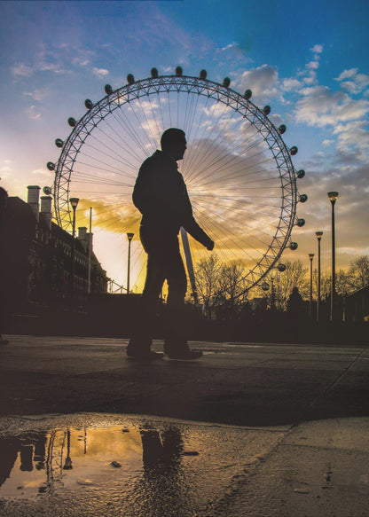 A low-angle shot of a person's silhouette walking past the London Eye at sunset, with the vibrant orange and blue sky reflected in a puddle in the foreground. Decor