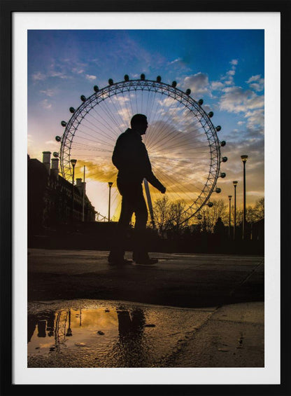 A low-angle shot of a person's silhouette walking past the London Eye at sunset, with the vibrant orange and blue sky reflected in a puddle in the foreground. Decor