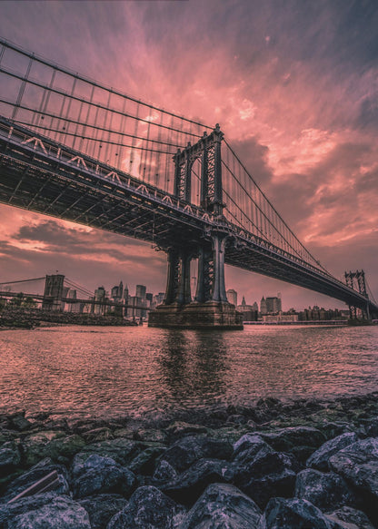 A dramatic low-angle view of the Manhattan Bridge over the East River at sunset. The sky is filled with pink and red clouds, which are reflected in the water below. The dark silhouette of the bridge contrasts with the vibrant sky, and the New York City skyline is visible in the background. In the foreground, dark, wet rocks line the riverbank. Decor
