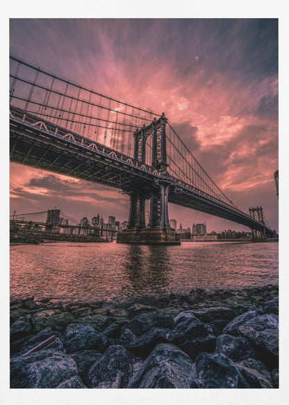 A dramatic low-angle view of the Manhattan Bridge over the East River at sunset. The sky is filled with pink and red clouds, which are reflected in the water below. The dark silhouette of the bridge contrasts with the vibrant sky, and the New York City skyline is visible in the background. In the foreground, dark, wet rocks line the riverbank. Decor