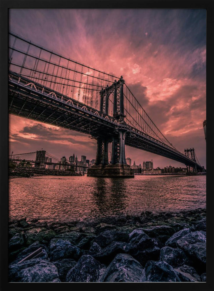 A dramatic low-angle view of the Manhattan Bridge over the East River at sunset. The sky is filled with pink and red clouds, which are reflected in the water below. The dark silhouette of the bridge contrasts with the vibrant sky, and the New York City skyline is visible in the background. In the foreground, dark, wet rocks line the riverbank. Decor