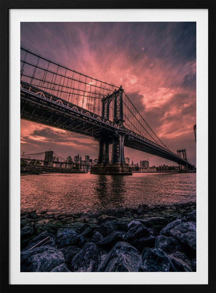 A dramatic low-angle view of the Manhattan Bridge over the East River at sunset. The sky is filled with pink and red clouds, which are reflected in the water below. The dark silhouette of the bridge contrasts with the vibrant sky, and the New York City skyline is visible in the background. In the foreground, dark, wet rocks line the riverbank. Decor