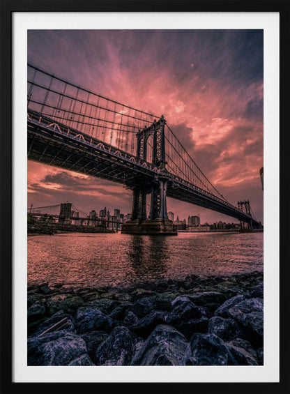 A dramatic low-angle view of the Manhattan Bridge over the East River at sunset. The sky is filled with pink and red clouds, which are reflected in the water below. The dark silhouette of the bridge contrasts with the vibrant sky, and the New York City skyline is visible in the background. In the foreground, dark, wet rocks line the riverbank. Decor