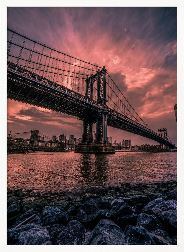 A dramatic low-angle view of the Manhattan Bridge over the East River at sunset. The sky is filled with pink and red clouds, which are reflected in the water below. The dark silhouette of the bridge contrasts with the vibrant sky, and the New York City skyline is visible in the background. In the foreground, dark, wet rocks line the riverbank. Decor