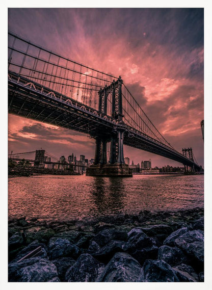 A dramatic low-angle view of the Manhattan Bridge over the East River at sunset. The sky is filled with pink and red clouds, which are reflected in the water below. The dark silhouette of the bridge contrasts with the vibrant sky, and the New York City skyline is visible in the background. In the foreground, dark, wet rocks line the riverbank. Decor