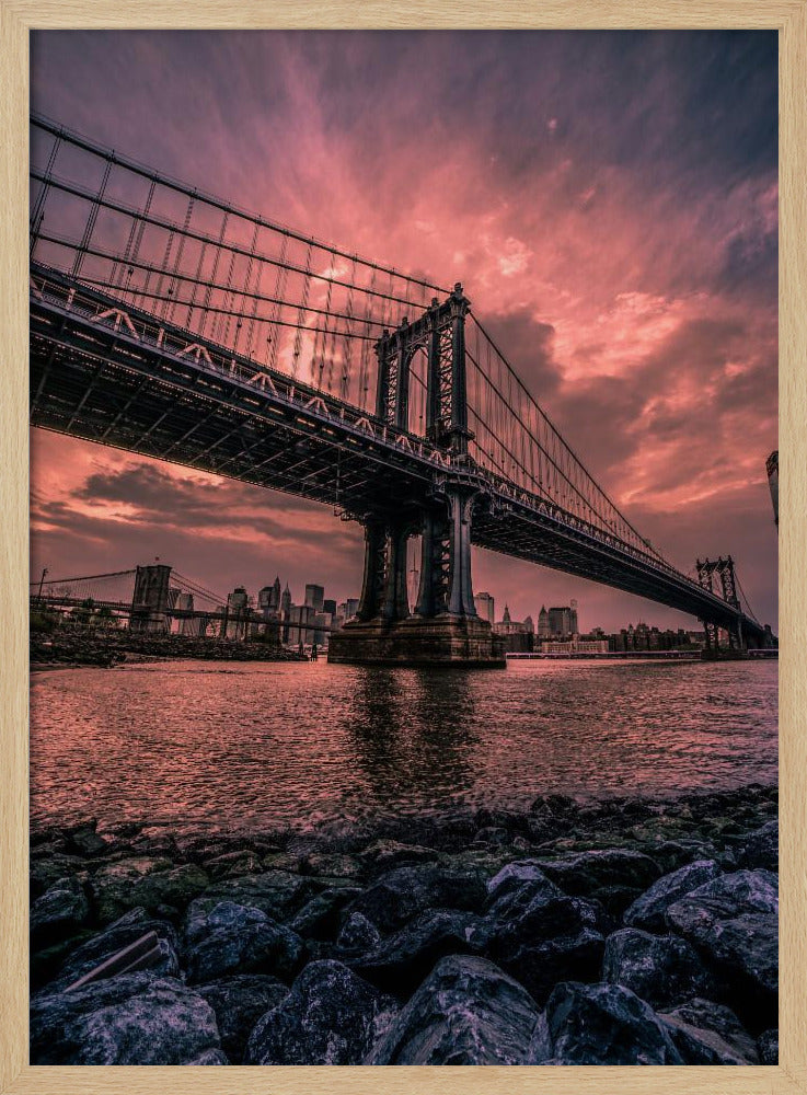 A dramatic low-angle view of the Manhattan Bridge over the East River at sunset. The sky is filled with pink and red clouds, which are reflected in the water below. The dark silhouette of the bridge contrasts with the vibrant sky, and the New York City skyline is visible in the background. In the foreground, dark, wet rocks line the riverbank. Decor