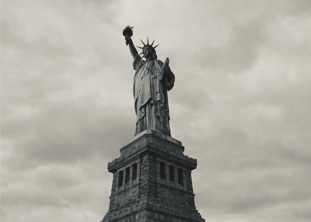 A low-angle, black and white photograph of the Statue of Liberty standing tall against a cloudy, dramatic sky, enclosed in a silver frame. Poster