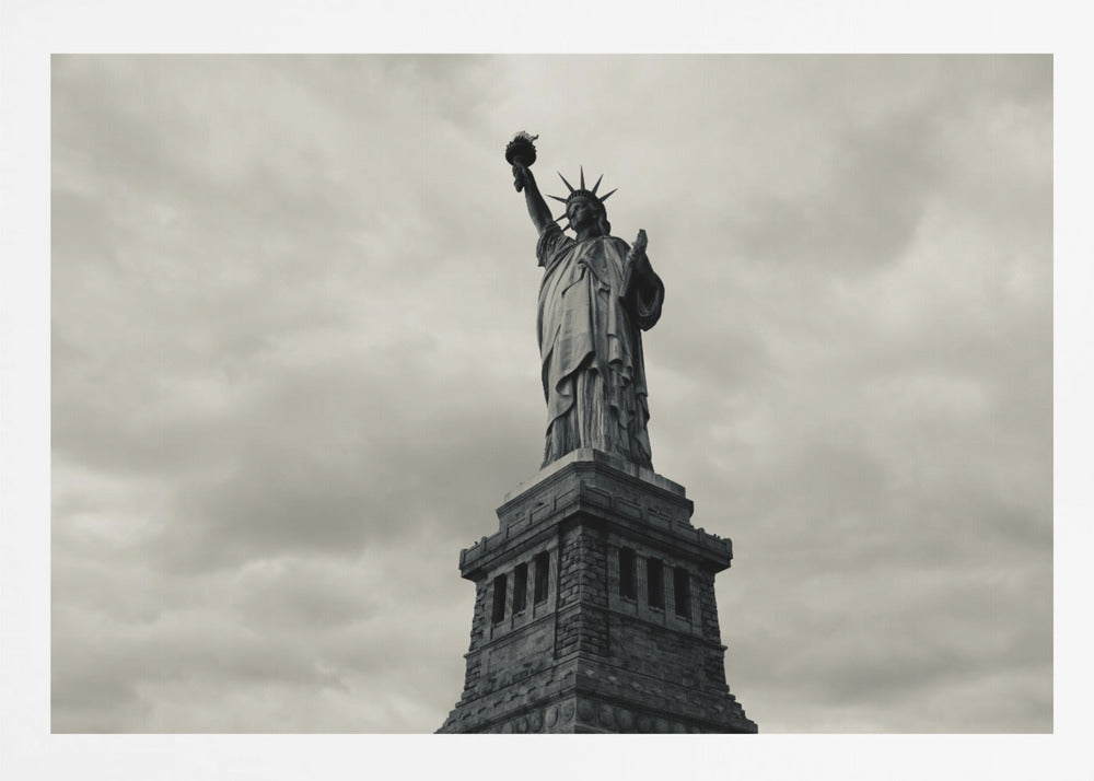 A low-angle, black and white photograph of the Statue of Liberty standing tall against a cloudy, dramatic sky, enclosed in a silver frame. Poster