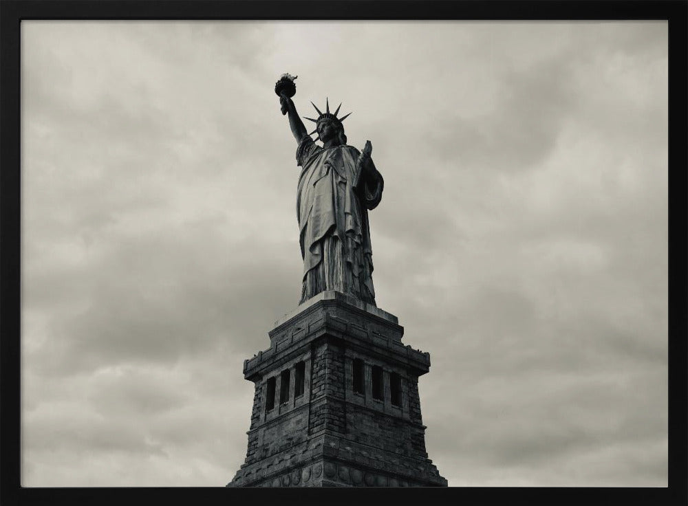 A low-angle, black and white photograph of the Statue of Liberty standing tall against a cloudy, dramatic sky, enclosed in a silver frame. Poster