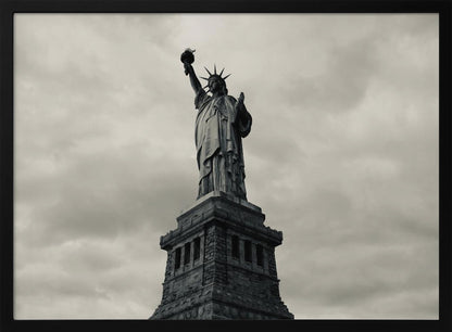 A low-angle, black and white photograph of the Statue of Liberty standing tall against a cloudy, dramatic sky, enclosed in a silver frame. Poster