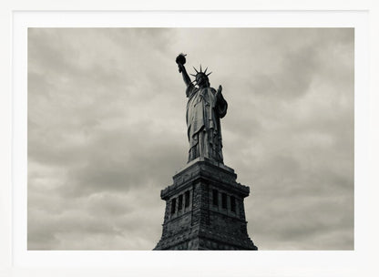 A low-angle, black and white photograph of the Statue of Liberty standing tall against a cloudy, dramatic sky, enclosed in a silver frame. Poster