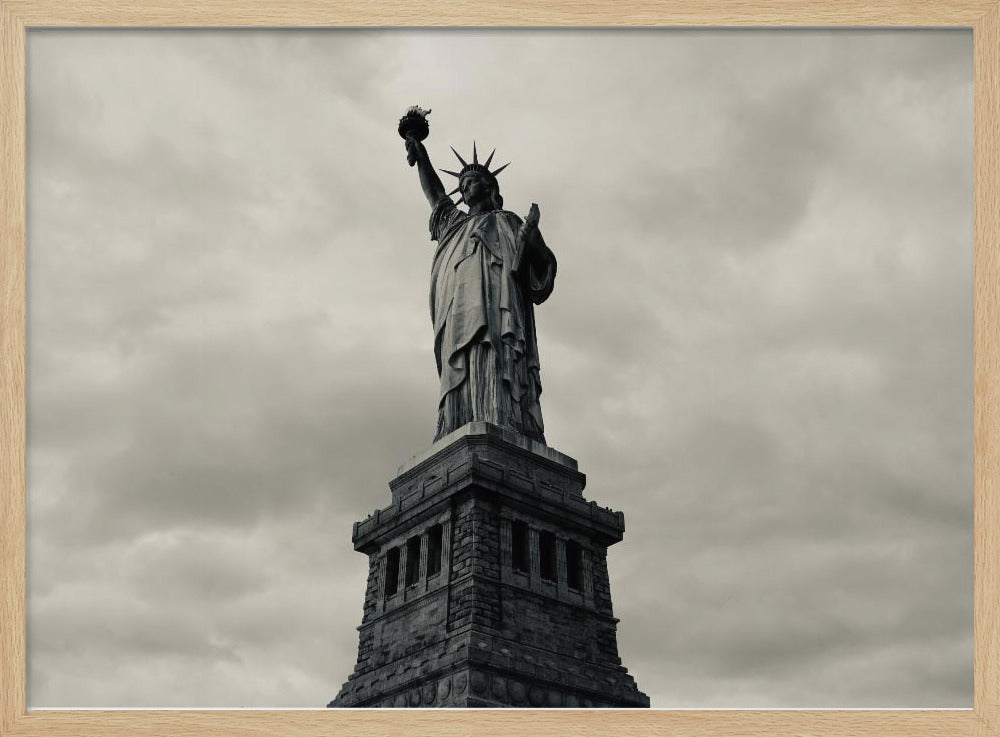 A low-angle, black and white photograph of the Statue of Liberty standing tall against a cloudy, dramatic sky, enclosed in a silver frame. Poster