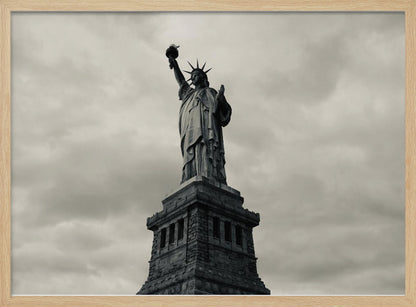A low-angle, black and white photograph of the Statue of Liberty standing tall against a cloudy, dramatic sky, enclosed in a silver frame. Poster