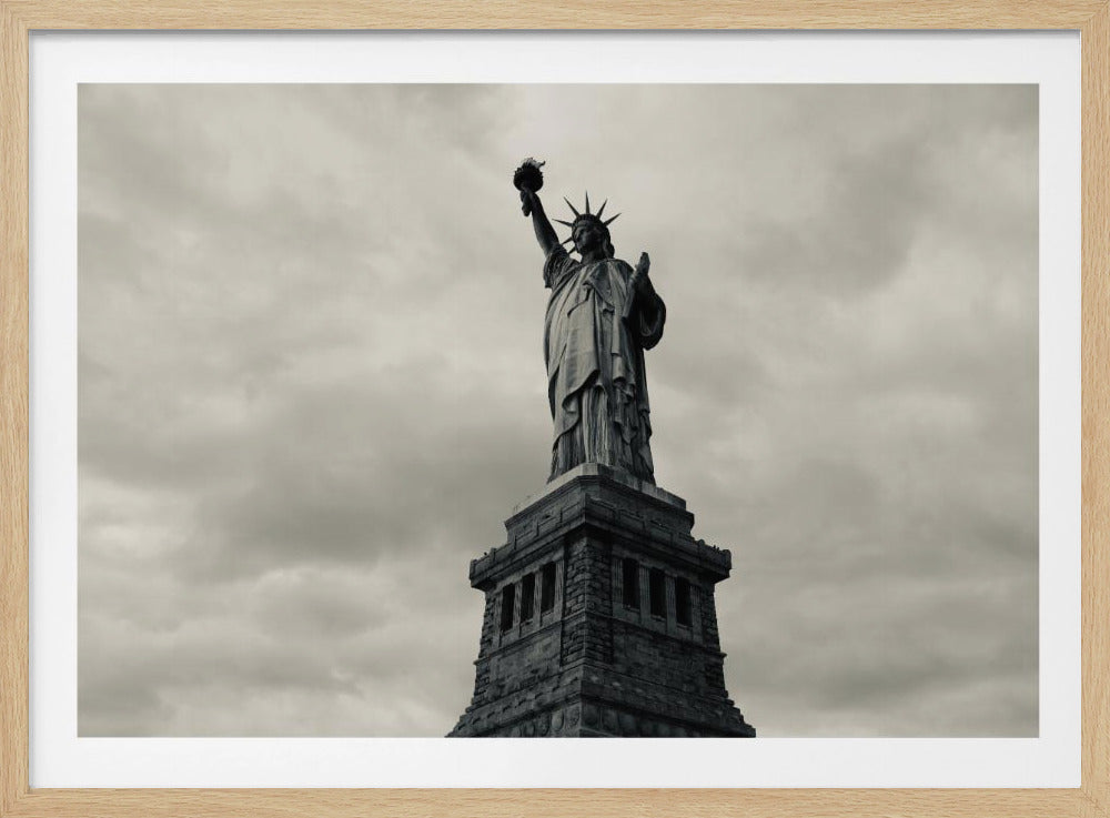 A low-angle, black and white photograph of the Statue of Liberty standing tall against a cloudy, dramatic sky, enclosed in a silver frame. Poster