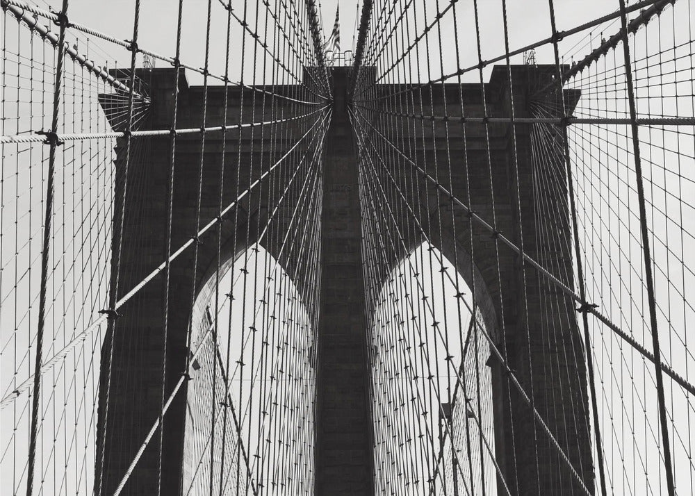 A low-angle, black and white photograph looking up at the iconic stone towers of the Brooklyn Bridge. The intricate web of suspension cables and vertical suspender ropes creates a symmetrical, geometric pattern against the light sky. The photo is enclosed in a silver frame. Print