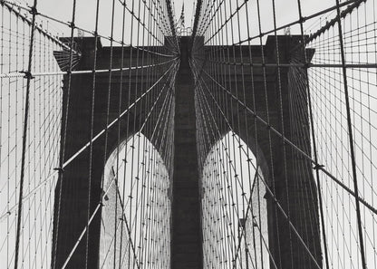 A low-angle, black and white photograph looking up at the iconic stone towers of the Brooklyn Bridge. The intricate web of suspension cables and vertical suspender ropes creates a symmetrical, geometric pattern against the light sky. The photo is enclosed in a silver frame. Print