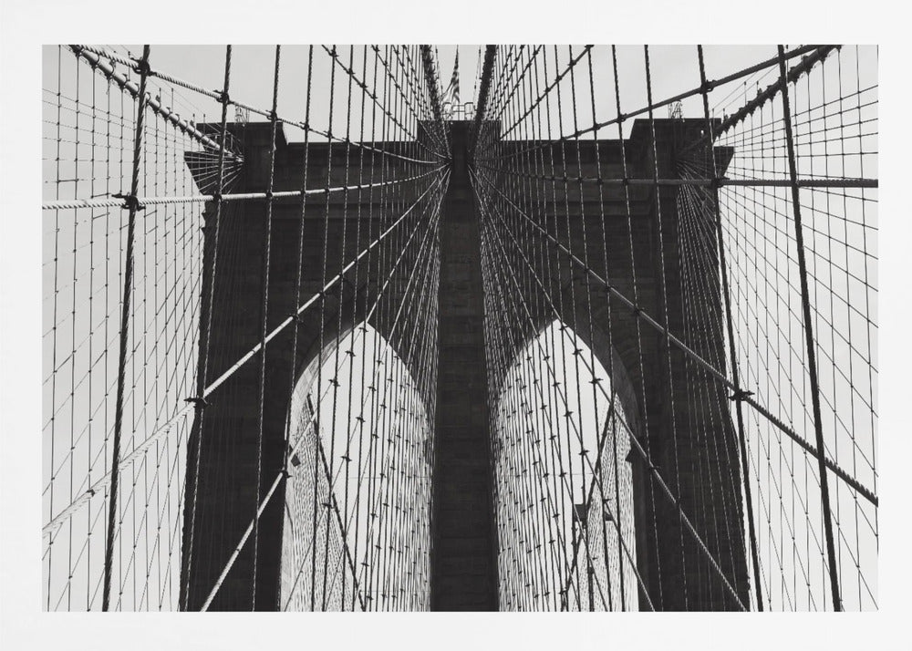 A low-angle, black and white photograph looking up at the iconic stone towers of the Brooklyn Bridge. The intricate web of suspension cables and vertical suspender ropes creates a symmetrical, geometric pattern against the light sky. The photo is enclosed in a silver frame. Print