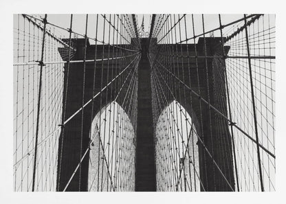 A low-angle, black and white photograph looking up at the iconic stone towers of the Brooklyn Bridge. The intricate web of suspension cables and vertical suspender ropes creates a symmetrical, geometric pattern against the light sky. The photo is enclosed in a silver frame. Print