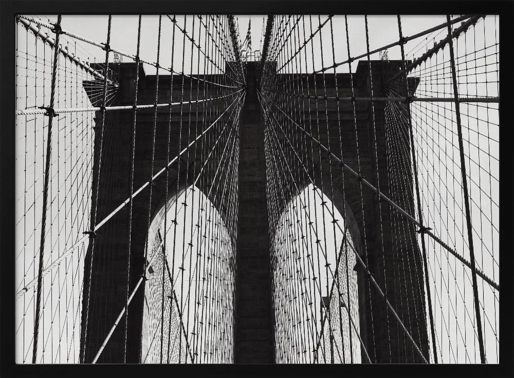 A low-angle, black and white photograph looking up at the iconic stone towers of the Brooklyn Bridge. The intricate web of suspension cables and vertical suspender ropes creates a symmetrical, geometric pattern against the light sky. The photo is enclosed in a silver frame. Print